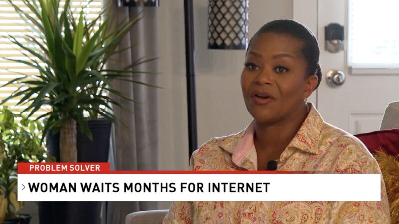 A woman sitting at home and talking to a news reporter. On screen, text says, 